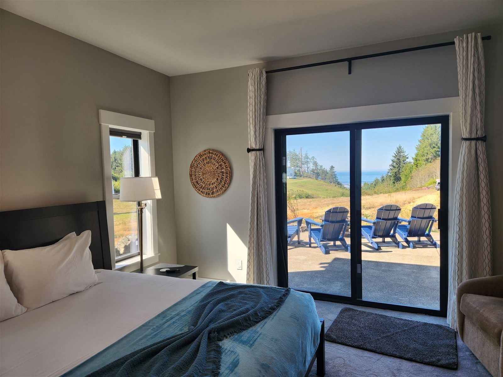 A hotel room with a bed, a desk, a lamp, a view of the ocean, and a balcony with chairs.