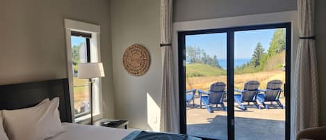 A hotel room with a bed, a desk, a lamp, a view of the ocean, and a balcony with chairs.