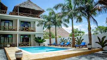A two-story building with a swimming pool, lounge chairs, and palm trees.
