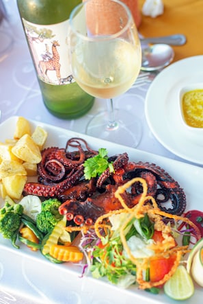 Ein Teller gegrillter Oktopus mit Gemüse und Kartoffeln, ein Glas Weißwein und eine Weinflasche mit Giraffendesign.