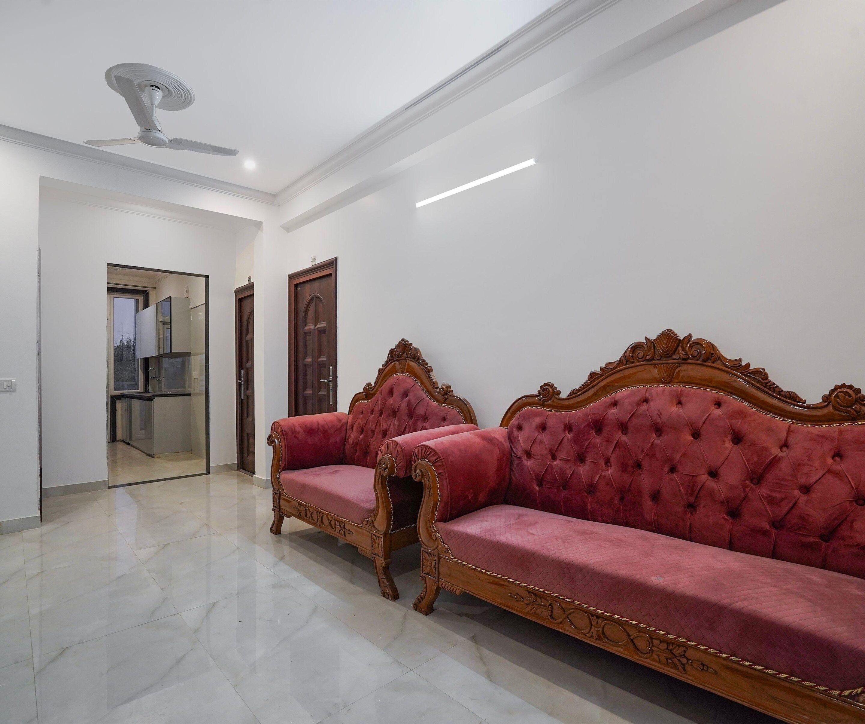 A spacious room with a large, ornate red sofa set against a white wall, a ceiling fan, and a door leading to another room.