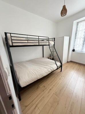 A bunk bed with a ladder, a small table, and a hanging light fixture in a room with wooden flooring and a window with curtains.