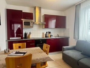 A compact kitchen with red cabinets, a microwave, and a stove. There is a dining table with chairs and a sofa in the living area. - Mountain View Studio "Hana" - Adler Resort (Kaprun)