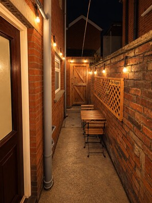 A narrow outdoor corridor with brick walls, wooden benches, and string lights. - Cosys Corner – Spacious 3-Bedroom Belfast Home (Belfast)