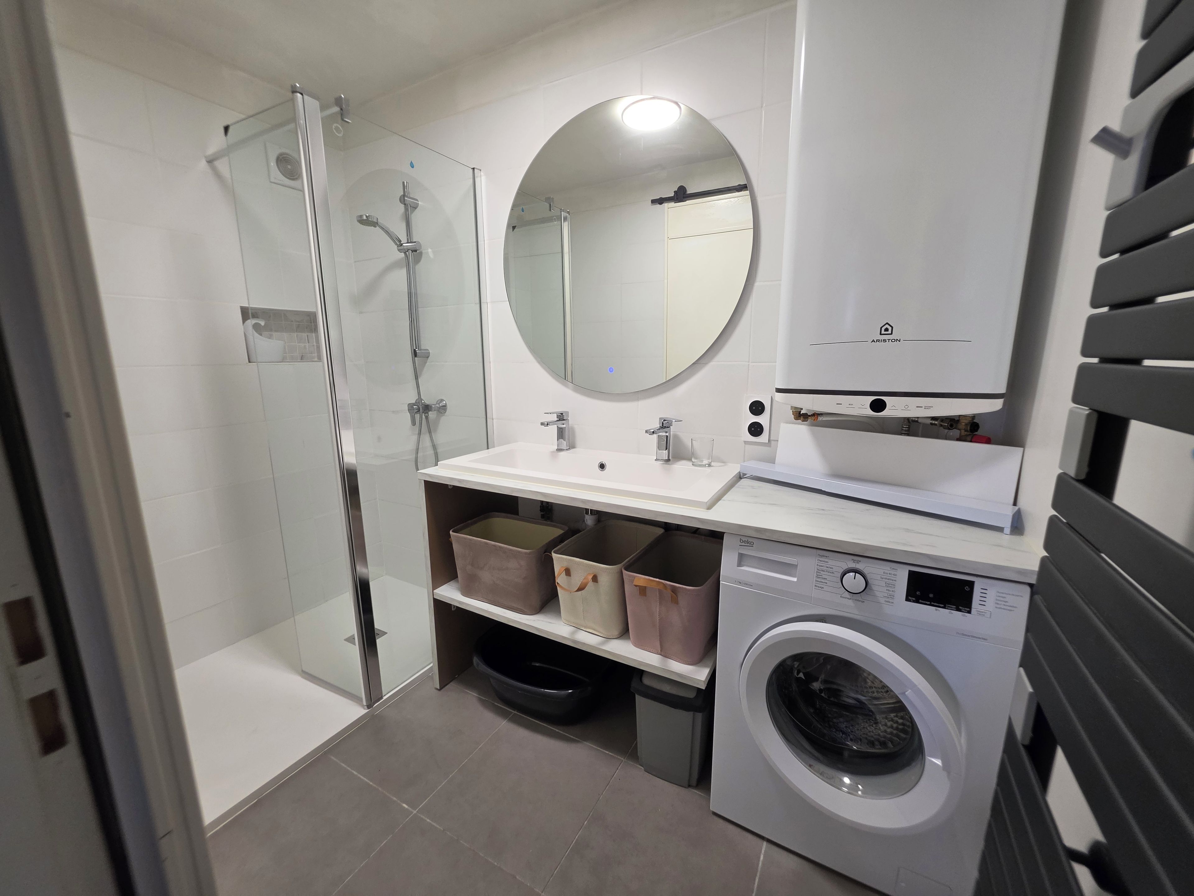 A modern bathroom with a glass-enclosed shower, a sink with a mirror above, a washing machine, and storage bins.