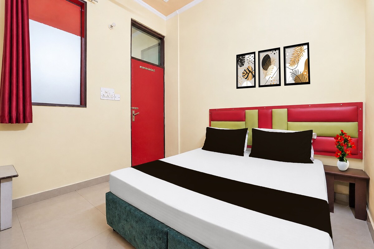 A hotel room with a bed, a red door, a nightstand, a vase with flowers, and three framed pictures on the wall.