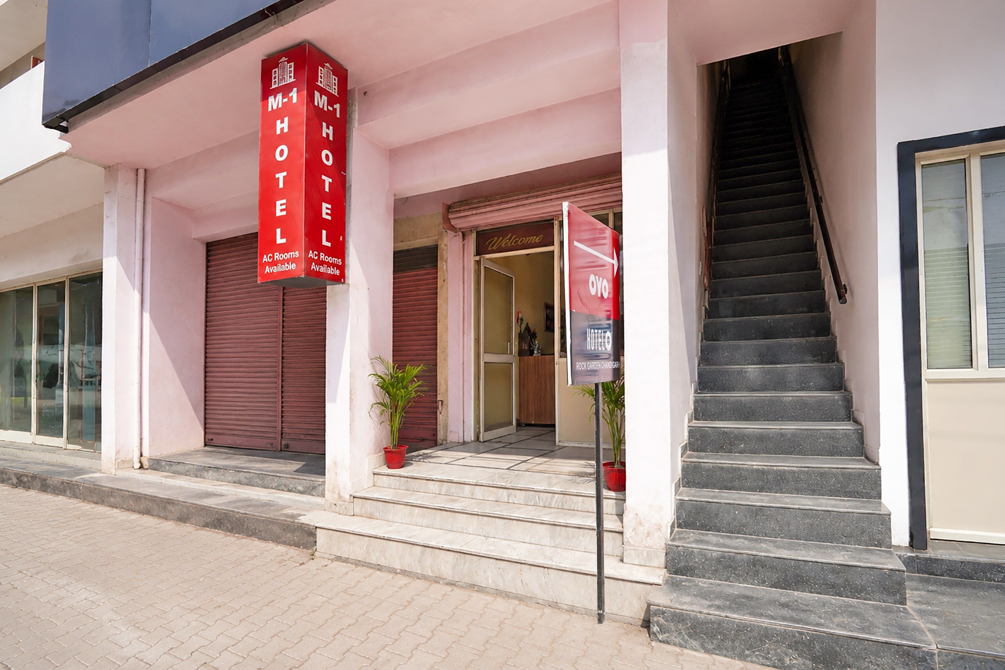 A hotel entrance with a staircase leading up to the door, a potted plant, and a red banner indicating AC rooms are available.