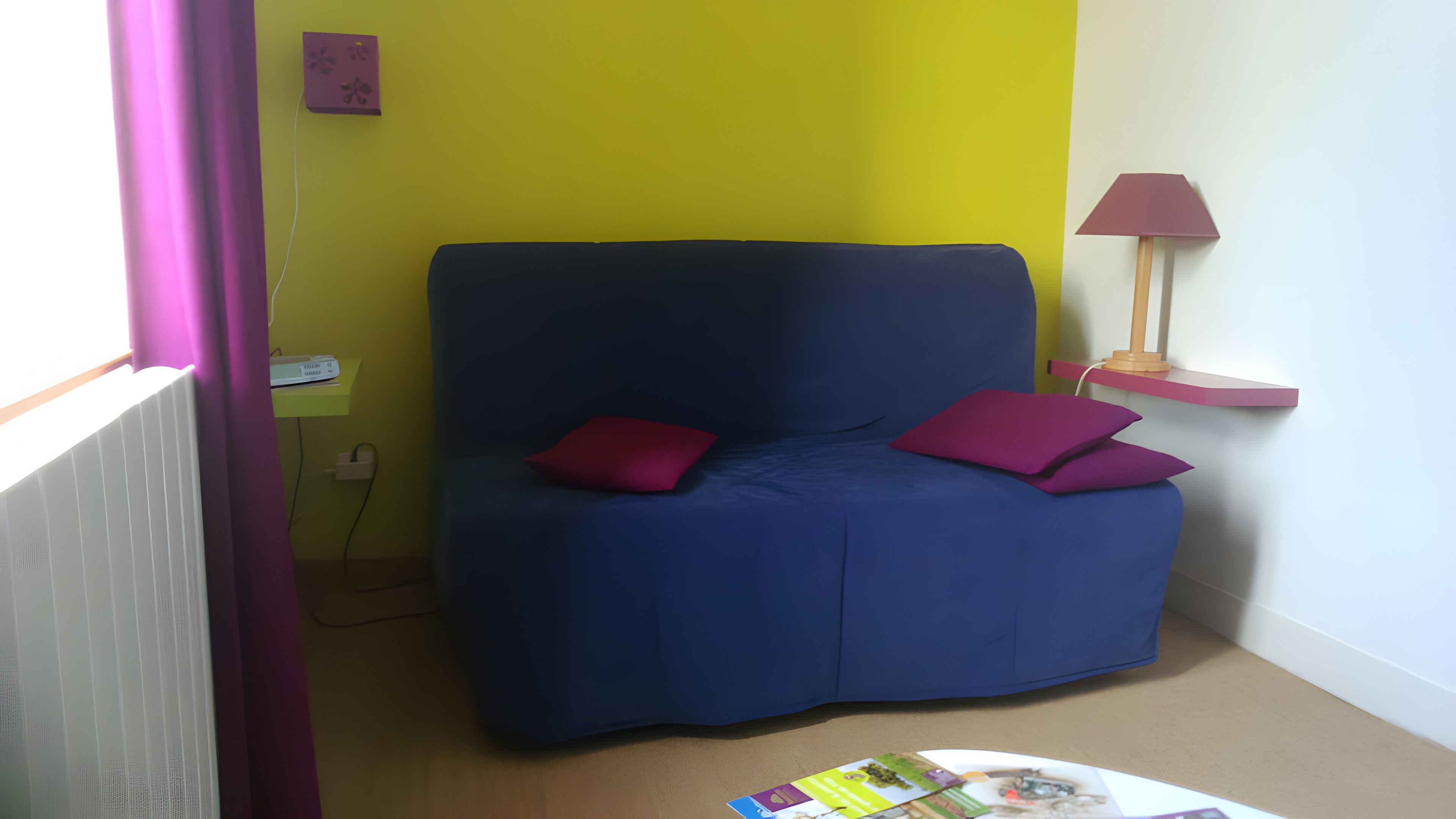 Een kamer met een blauwe bank, twee rode kussens, een tafeltje met tijdschriften, een lamp en een geel accent op de muur.