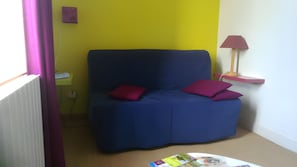 A room with a blue sofa, two red cushions, a small table with magazines, a lamp, and a wall with a yellow accent.