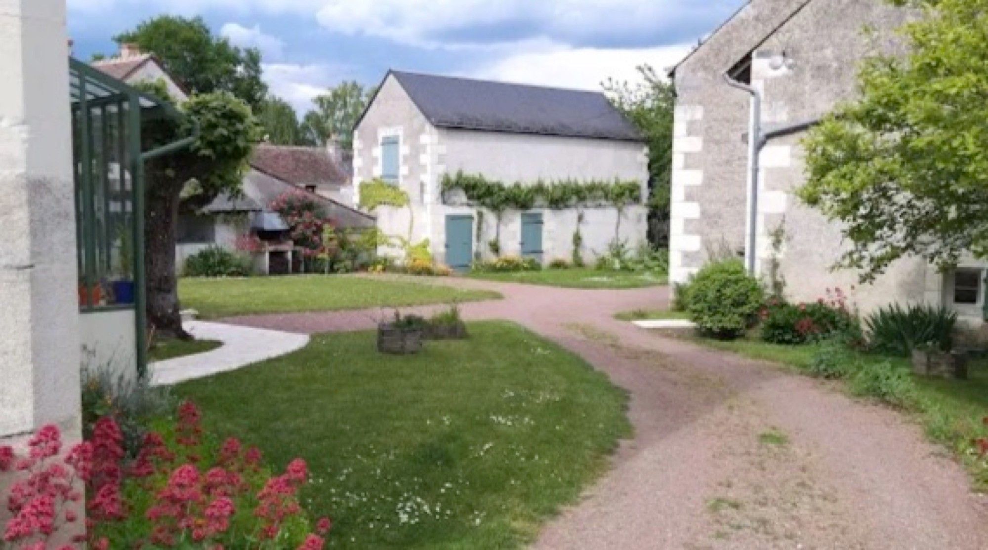 A residential property with a paved driveway, a garden with flowers, and a house with a grey roof and white walls.