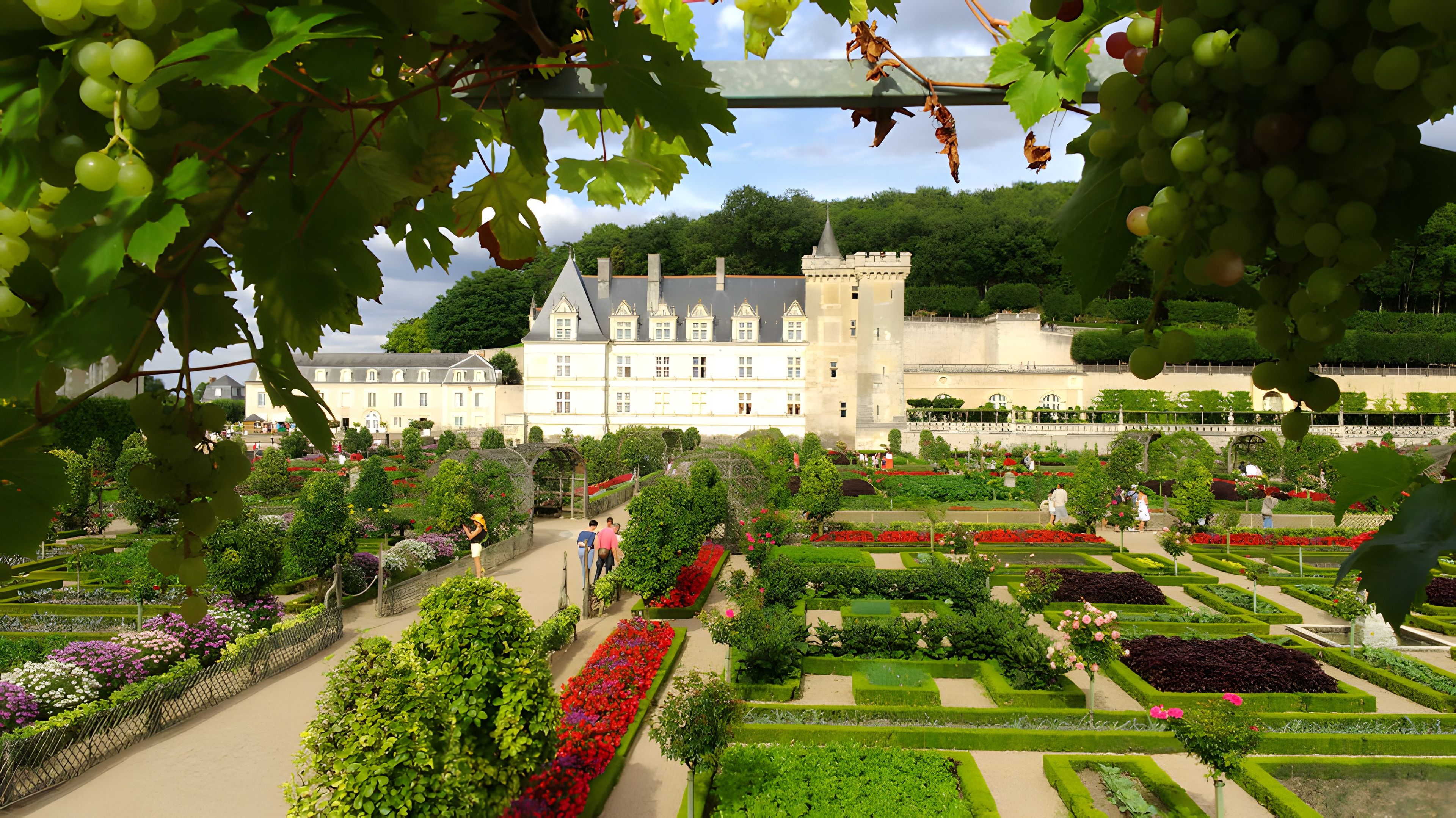 Un giardino formale con siepi curate e aiuole fiorite, un grande edificio con una torre e visitatori che passeggiano lungo i sentieri.