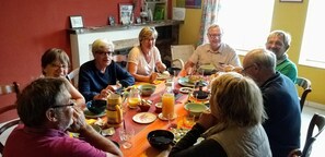 A group of people seated around a table with various dishes and drinks, in a room with a fireplace and a shelf with items.