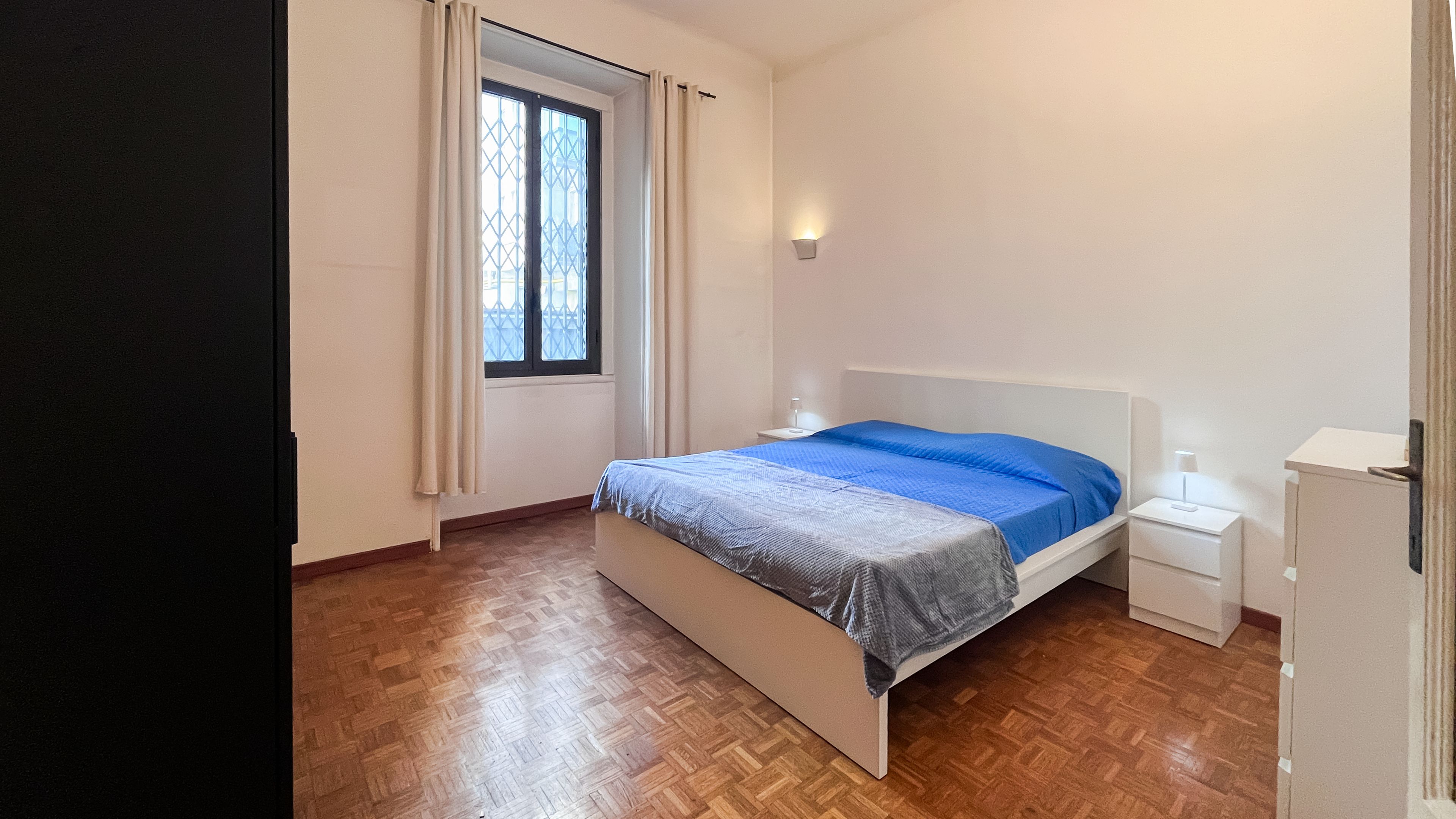 A bedroom with a bed, a nightstand, a bedside table, a window with curtains, and a wall-mounted light.