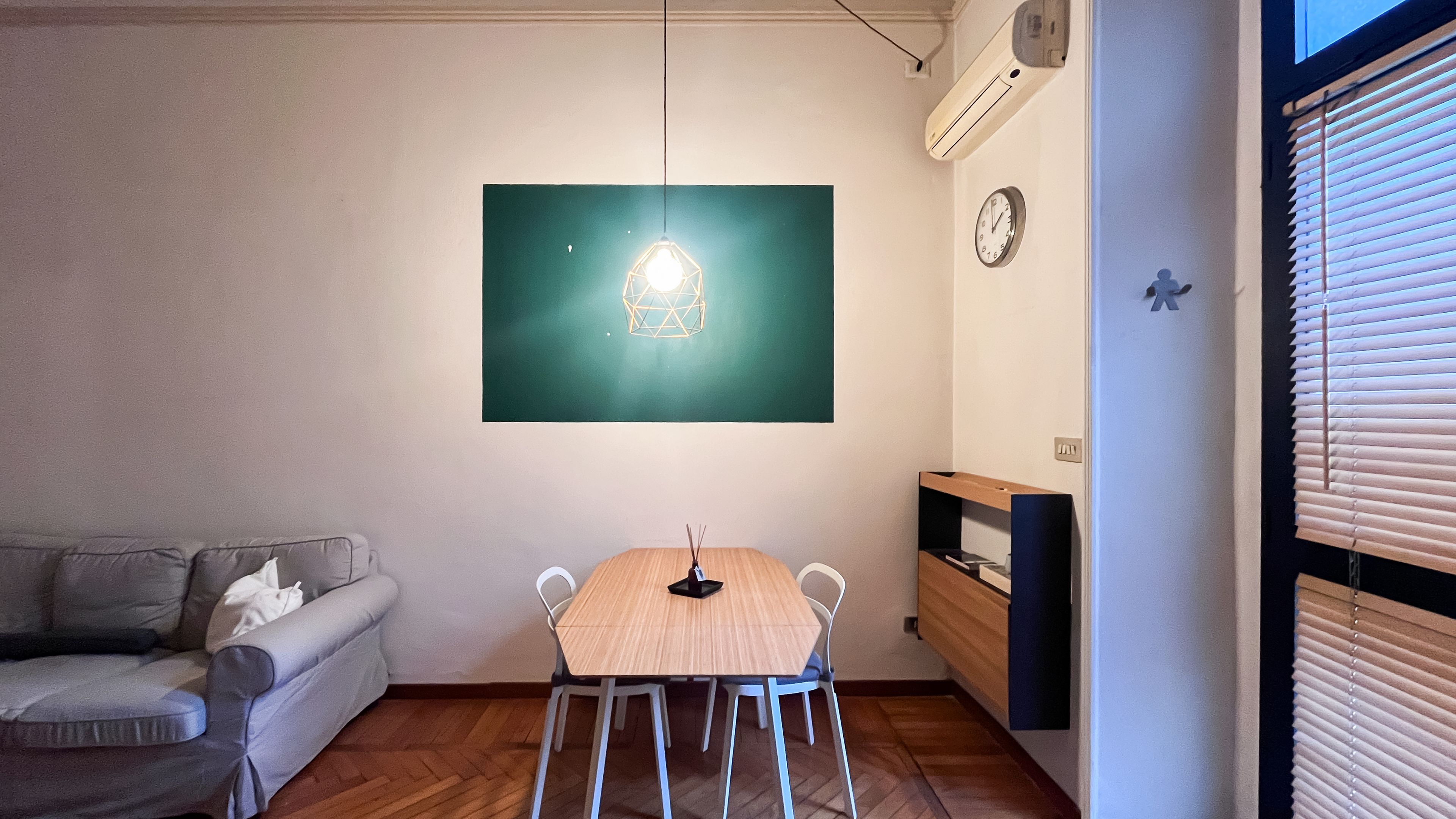 A room with a wooden dining table, a white chair, a grey sofa, a wall-mounted air conditioner, and a clock.