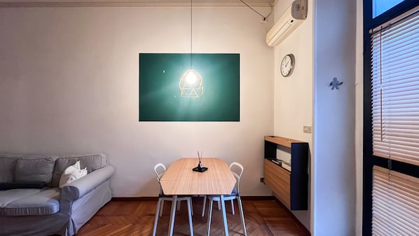 A room with a wooden dining table, a white chair, a grey sofa, a wall-mounted air conditioner, and a clock. - Cambiasi Apartment (Milano)