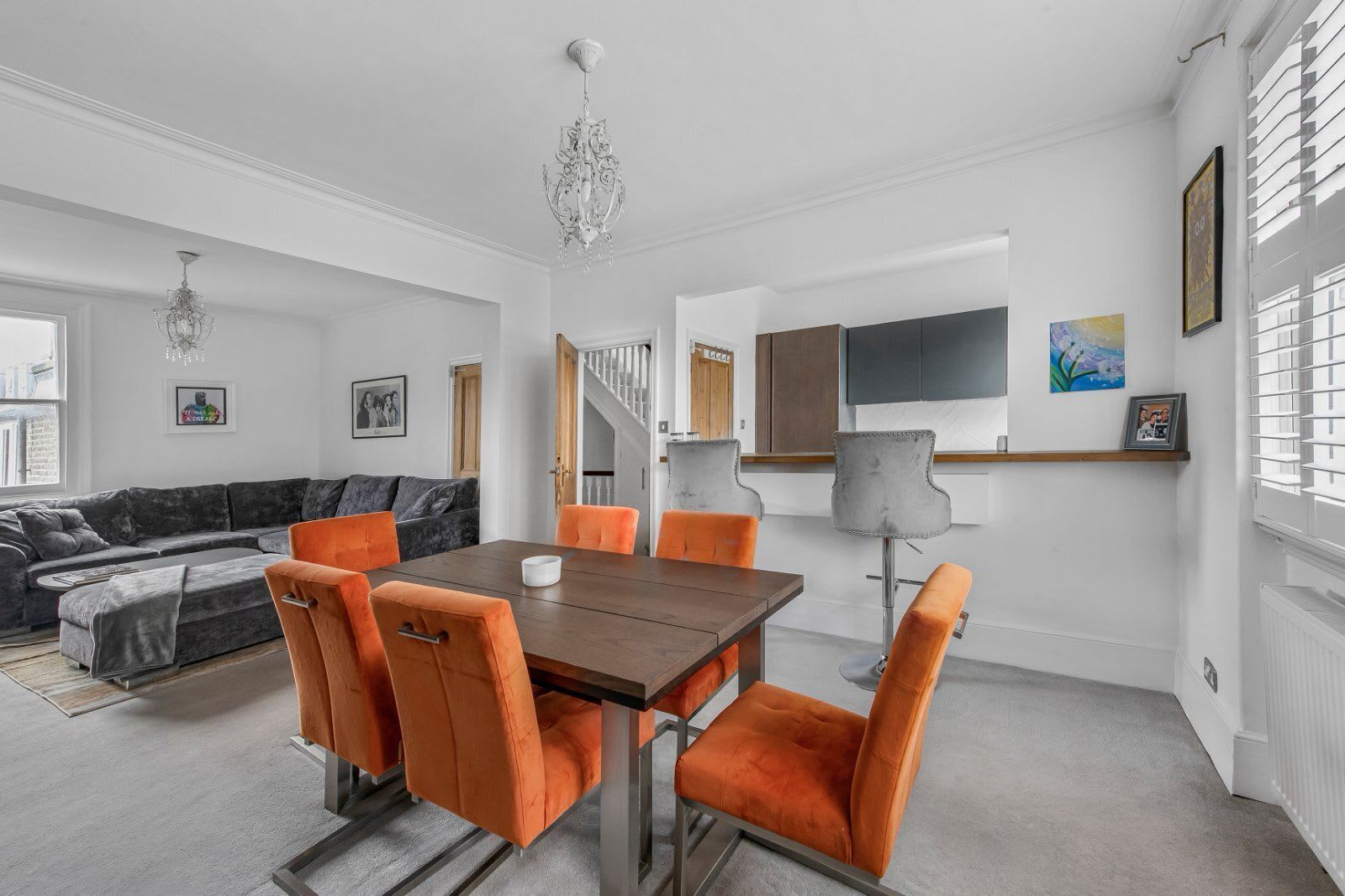 Een moderne woonkamer met een grijze bank, oranje fauteuils, een houten eettafel en een kroonluchter.