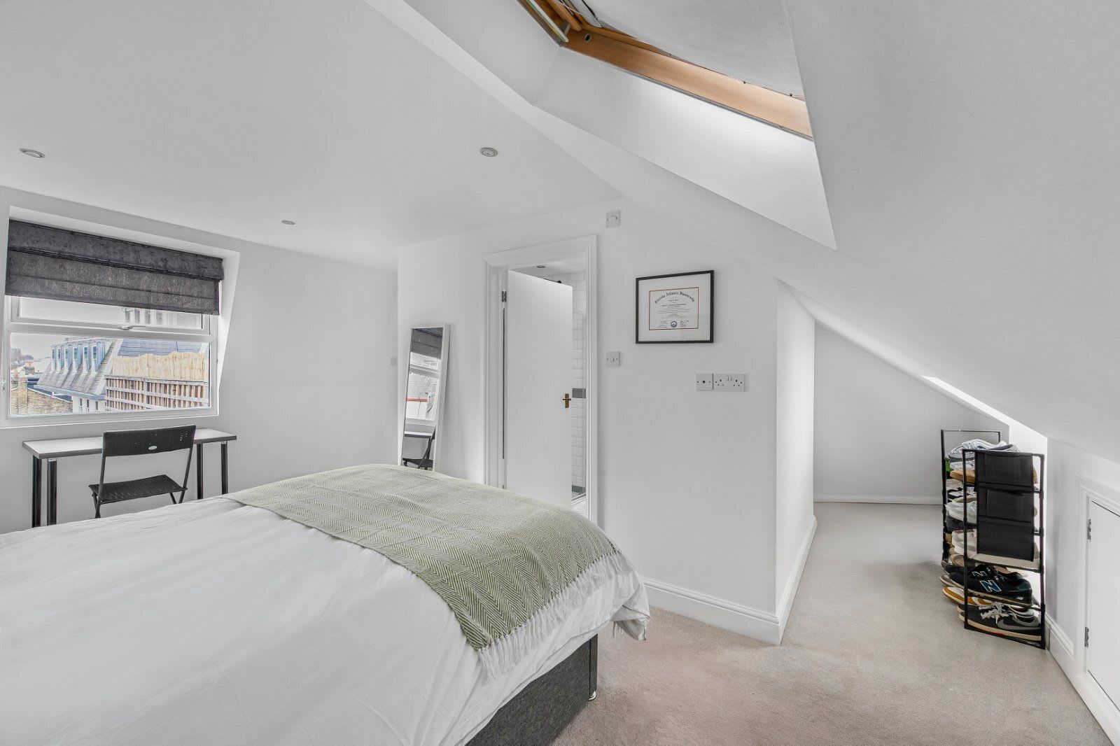 Een slaapkamer met een bed, bureau, stoel, raam met uitzicht, deur en een schoenenrek.