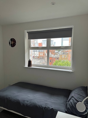 A single bed with a dark mattress and pillow, a window with a view of buildings, and a wall-mounted object.