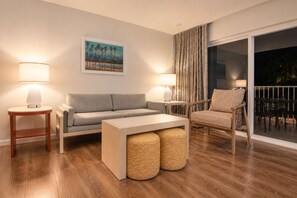 A modern living room with a sofa, two side tables, a chair, and ottomans. There is a painting on the wall and a view of the outdoors through a window. - 2BR Apt near Kam 1 Beach, Pool, Hot Tub, BBQ (Kihei)