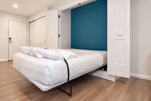 A bedroom with a bed, white walls, a blue accent wall, and wooden flooring. - 2BR Apt near Kam 1 Beach, Pool, Hot Tub, BBQ (Kihei)
