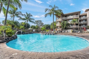 A swimming pool with a clear blue water, surrounded by palm trees and a building with balconies. - 2BR Apt near Kam 1 Beach, Pool, Hot Tub, BBQ (Kihei)
