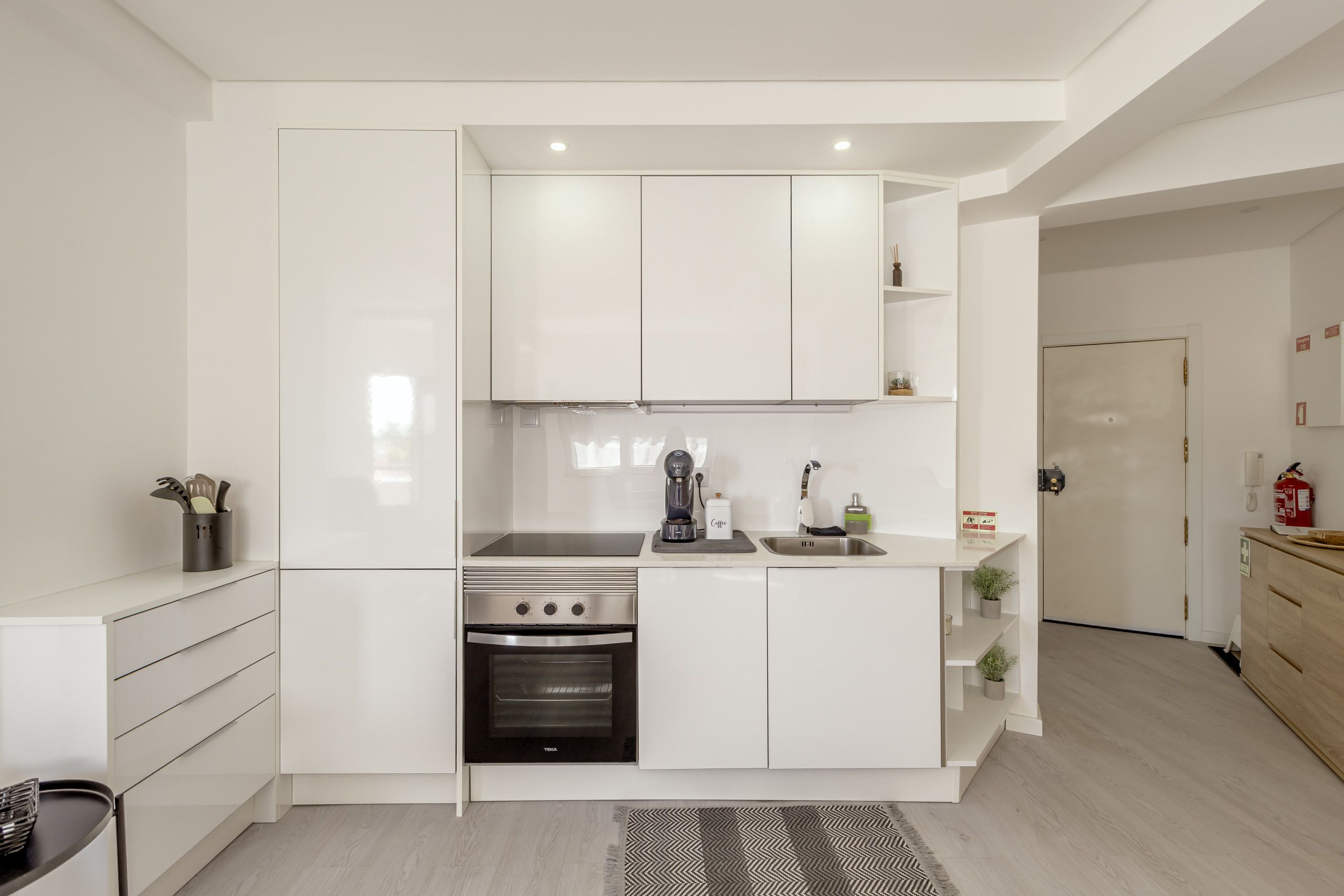 Una cocina moderna con muebles blancos, horno empotrado, cafetera y pileta de acero inoxidable.