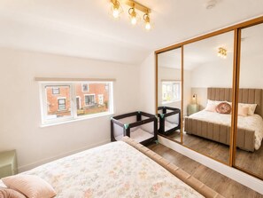 A bedroom with a large mirror, a bed with floral bedding, a window with a view of a brick building, and a small enclosed play area. - The Cosy Claddagh – Spacious 3-Bedroom Belfast Home (Belfast)