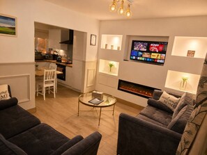 Ein modernes Wohnzimmer mit Kamin, einem an der Wand montierten Fernseher und einem durch einen offenen Durchgang einsehbaren Essbereich.