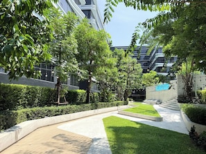 A landscaped outdoor area with pathways, greenery, and modern buildings in the background. - Serene 1-bedroom Apartment With Pool & Gym in Khlong Toei (Bangkok)