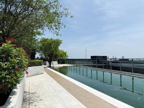 A rooftop pool with a glass railing, surrounded by greenery and a city skyline in the background. - Serene 1-bedroom Apartment With Pool & Gym in Khlong Toei (Bangkok)