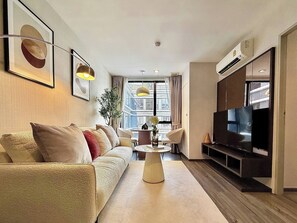 A modern living room with a sofa, a coffee table, a TV, and a large window with a city view. - Serene 1-bedroom Apartment With Pool & Gym in Khlong Toei (Bangkok)