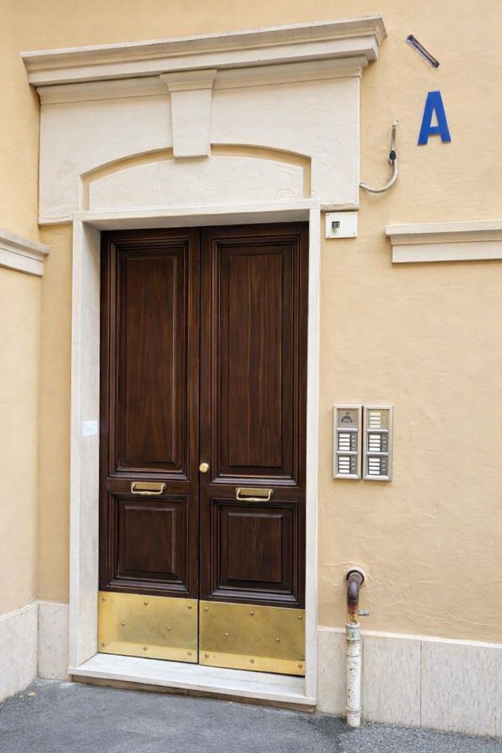 Una doble puerta de madera con manijas de latón y un umbral de bronce, ubicada en una pared beige con un letrero azul con la letra 'A'.