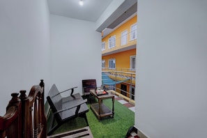 A small, enclosed outdoor area with a wooden bench, a table, and a chair. There is a balcony with a railing and a view of a yellow building.