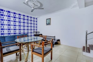A dining area with a round glass table, wooden chairs, a blue and white patterned wall, a ceiling fan, and a small framed picture on the wall.