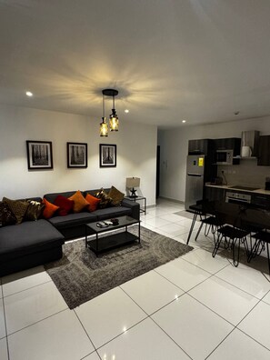 A modern living room with a sectional sofa, a coffee table, and a dining area with chairs. There is a kitchen with a refrigerator and microwave. - Luxury 3BR Apt w/ Pool, Gym Restaurant & DSTV - Embassy Gardens / 2 min from KIA (Accra)