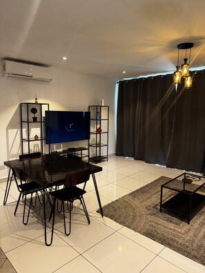 A modern living room with a flat-screen TV, a dining table, chairs, a coffee table, and a large window with curtains. - Luxury 3BR Apt w/ Pool, Gym Restaurant & DSTV - Embassy Gardens / 2 min from KIA (Accra)