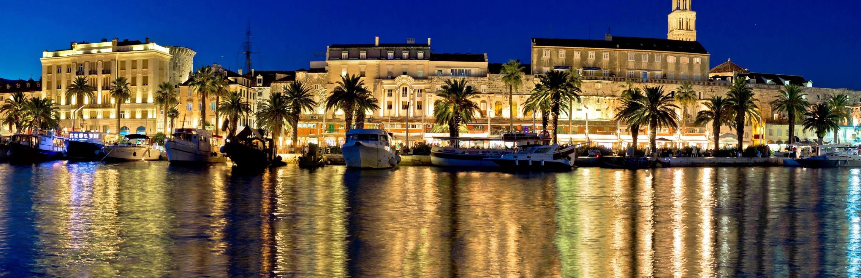 Een jachthaven aan het water met aangemeerde boten, gebouwen in klassieke stijl en palmbomen langs de boulevard.