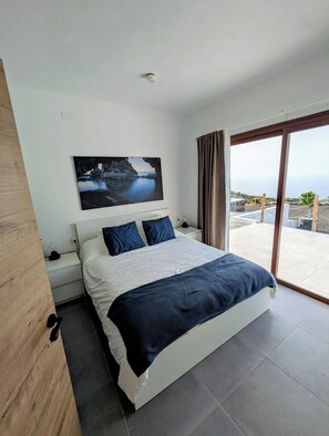 A modern bedroom with a large bed, a nightstand, a wall-mounted picture, and a view of the outdoors through a sliding glass door. - Ensueño Estate (Frontera)