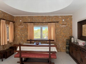 A bedroom with a stone wall, a bed with a wooden headboard, a bench, a window with curtains, and a dresser with decorative items. - Villa with heatable pool in Carvoeiro (Carvoeiro)