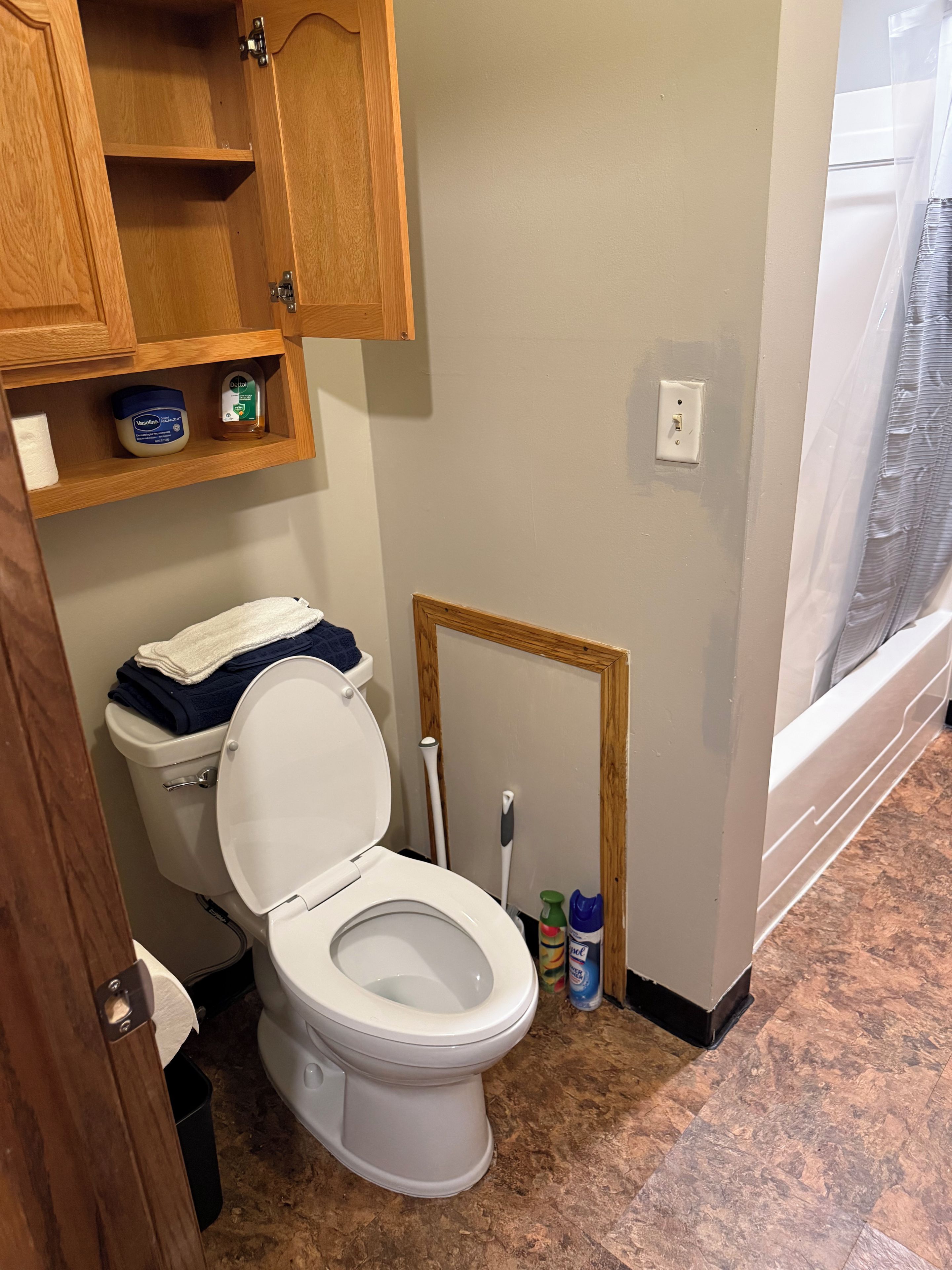 A bathroom with a toilet, wooden cabinets, a shelf with toiletries, and a window with a view.