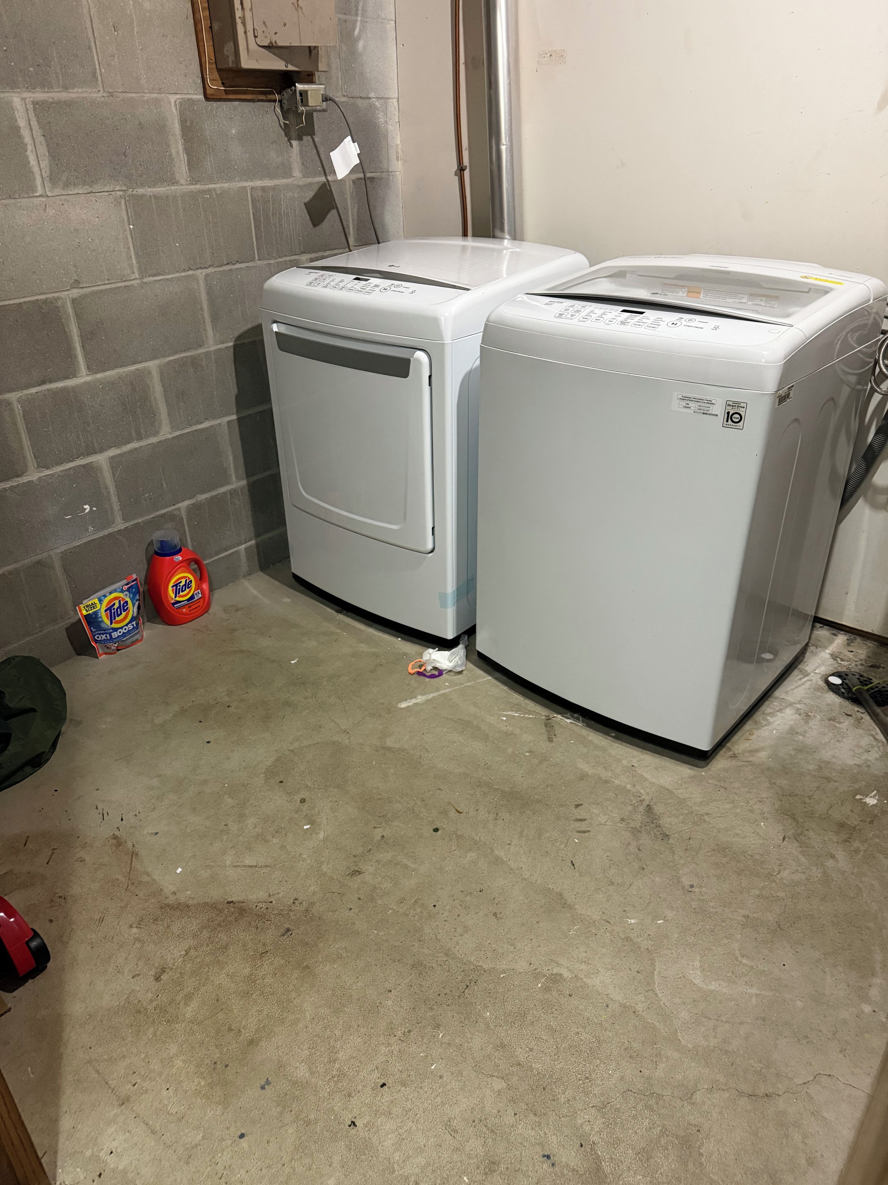 Two white front-loading washing machines in a laundry room with a concrete floor, a brick wall, and a metal vent.