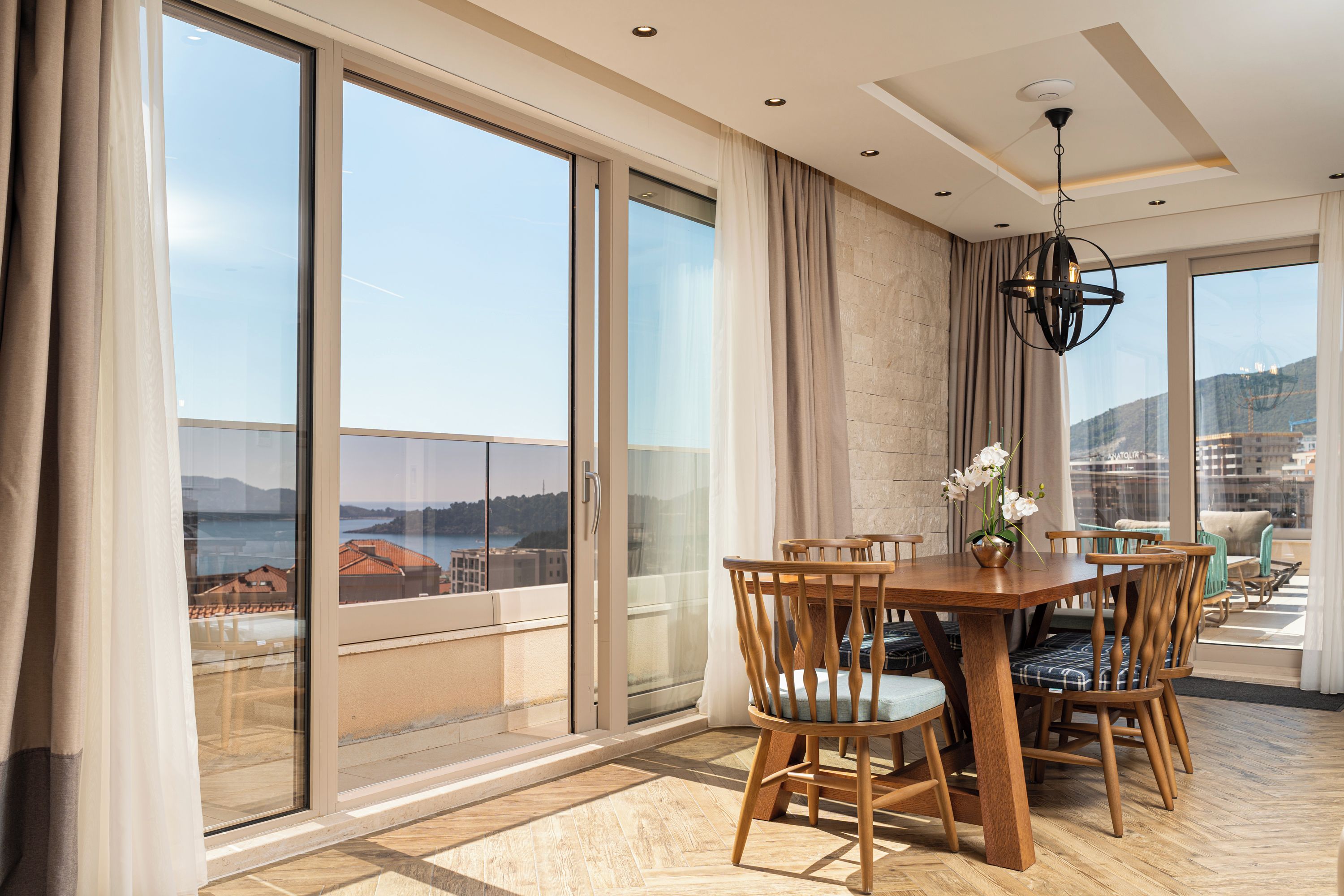 A dining area with a wooden table and chairs, large windows with curtains, and a view of the sea.