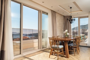 A dining area with a wooden table and chairs, large windows with curtains, and a view of the sea. - Garni Casa Di Sofia (Rafajlovići)