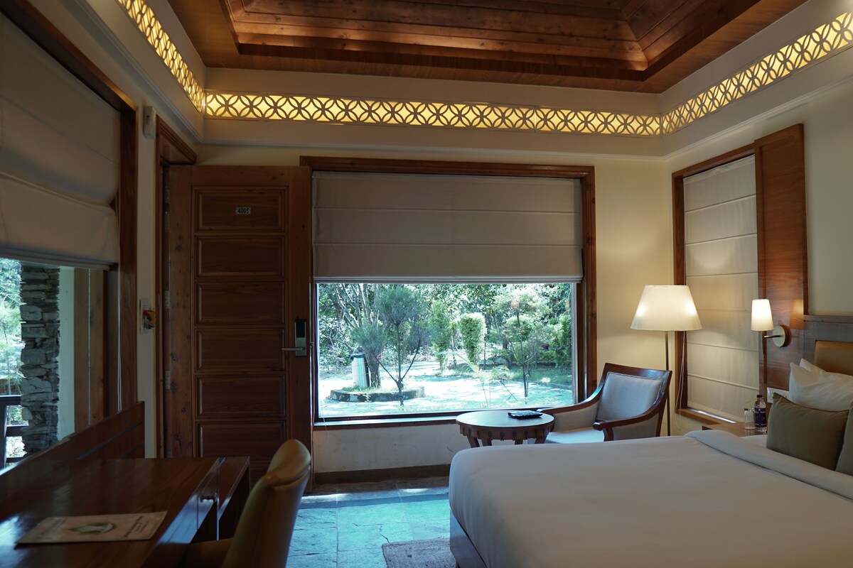 A hotel room with a bed, a desk, a chair, a window with a view of trees, and a decorative ceiling light fixture.