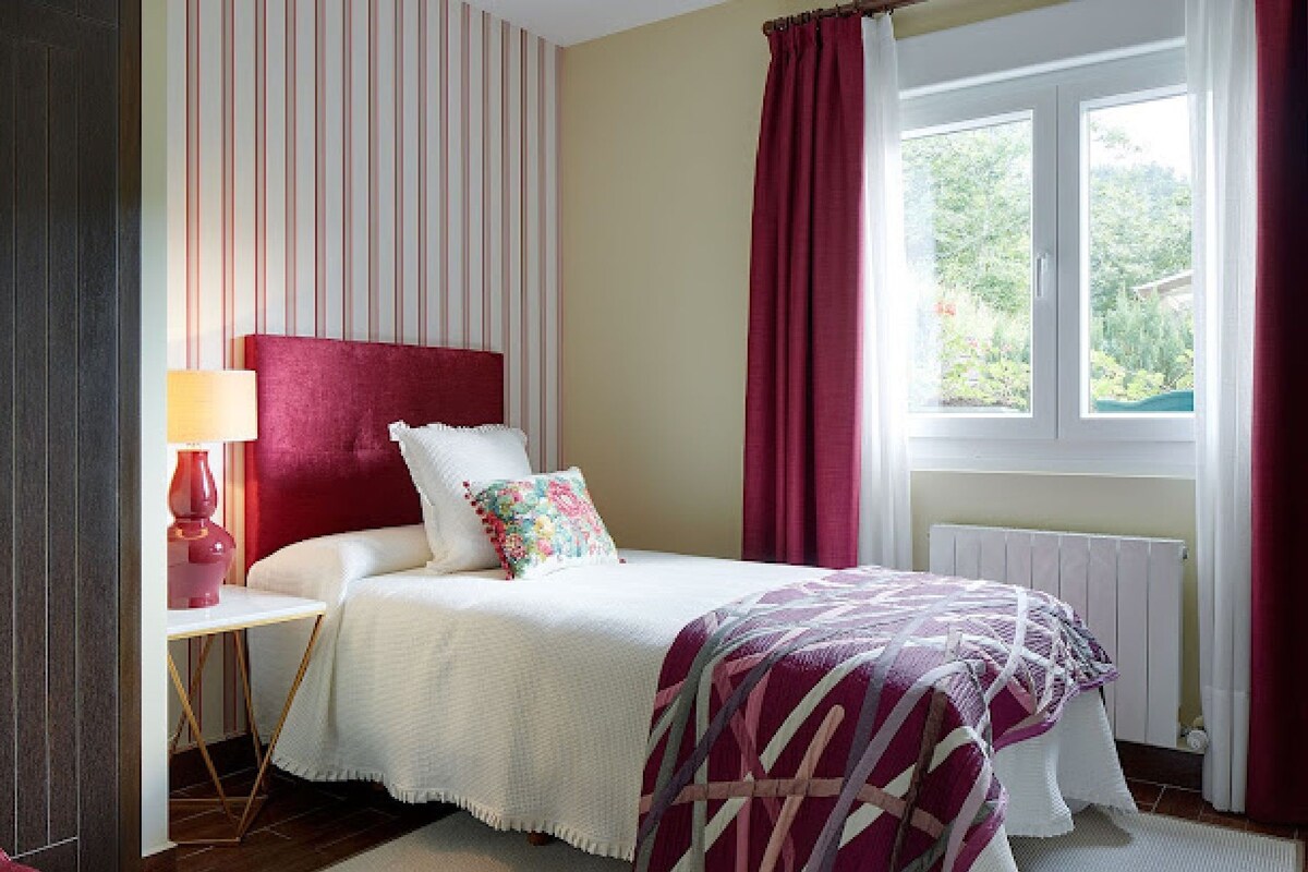 A bedroom with a bed, a nightstand, a lamp, a window with curtains, and a radiator.