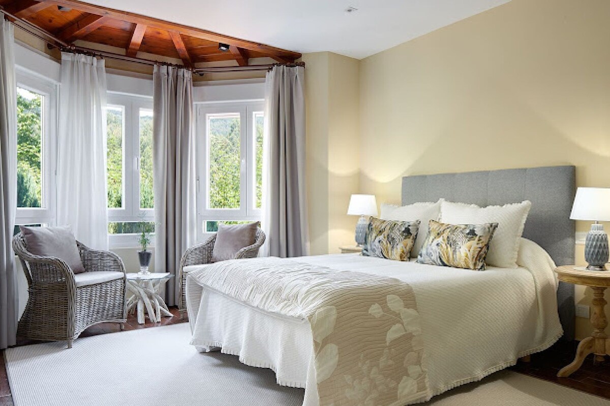 A bedroom with a large bed, two bedside tables with lamps, a chair, and a view of greenery through the windows.
