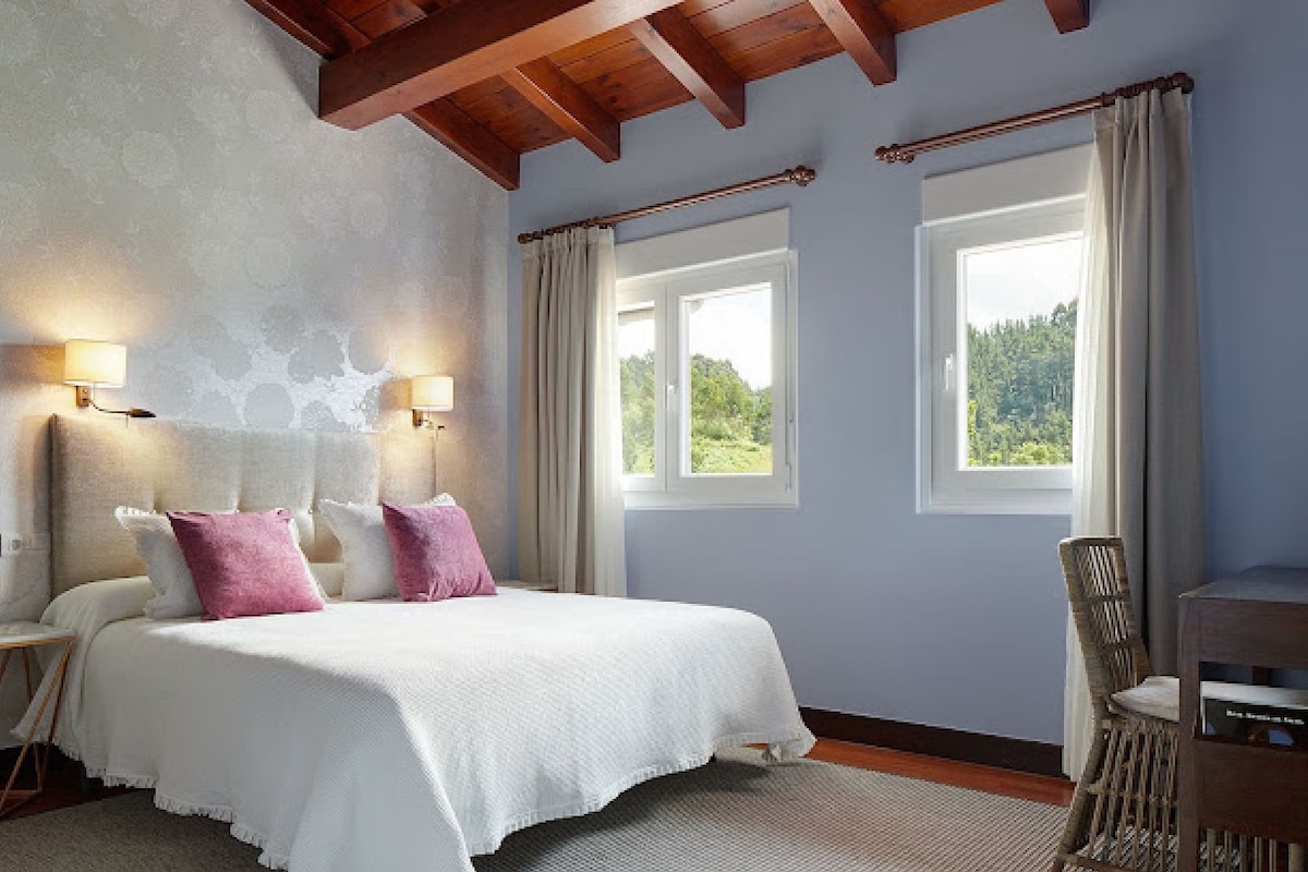 A bedroom with a bed, two bedside lamps, a chair, and two windows with curtains.