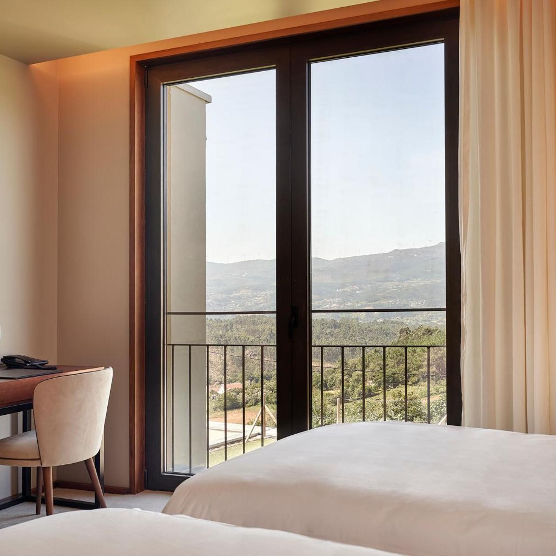 A hotel room with a bed, a desk, and a view of a mountainous landscape through a large window.