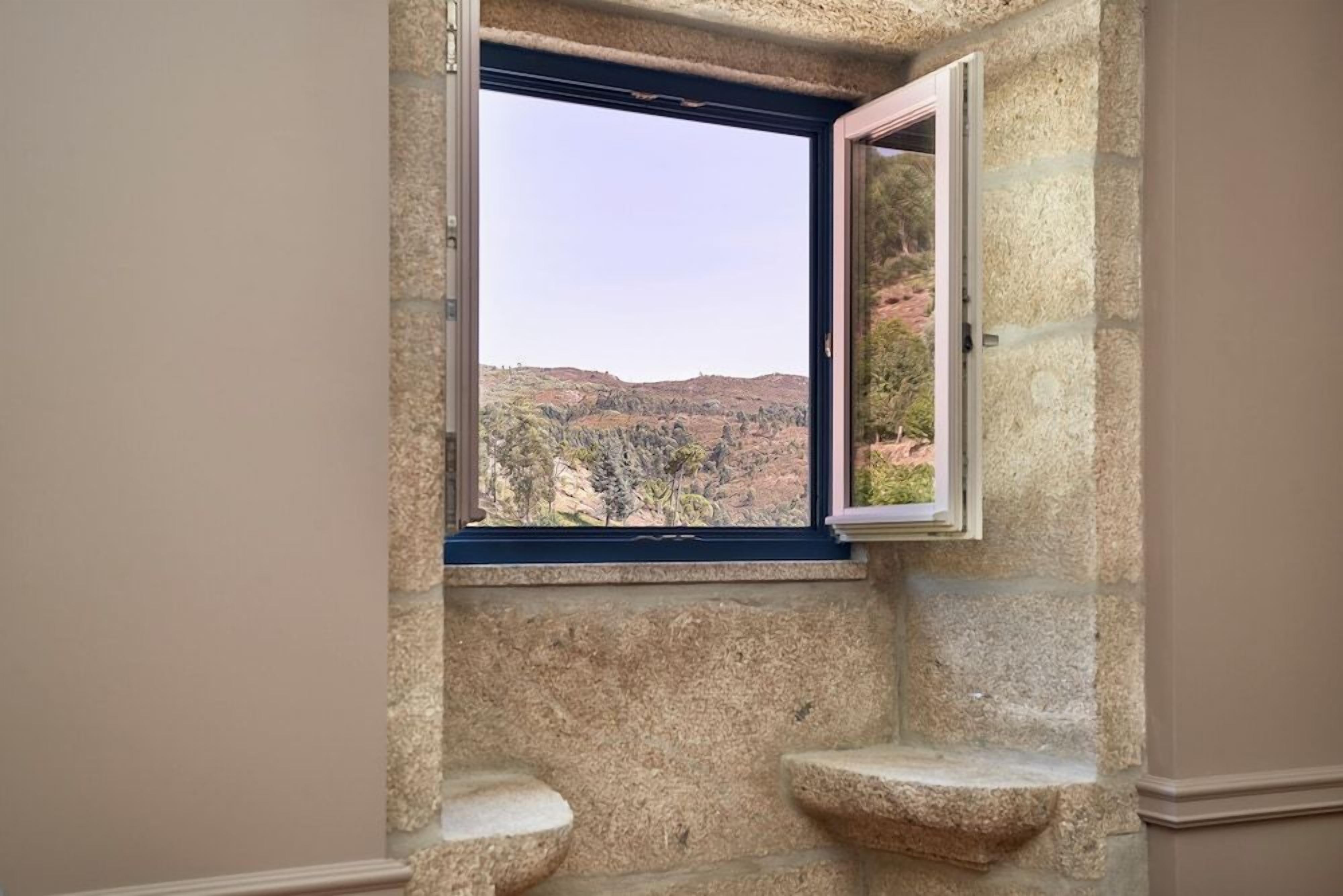 A bathroom with a stone wall, a window with an open view of a hilly landscape, and a small sink.