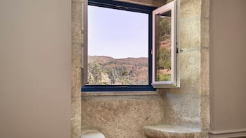 A bathroom with a stone wall, a window with an open view of a hilly landscape, and a small sink.
